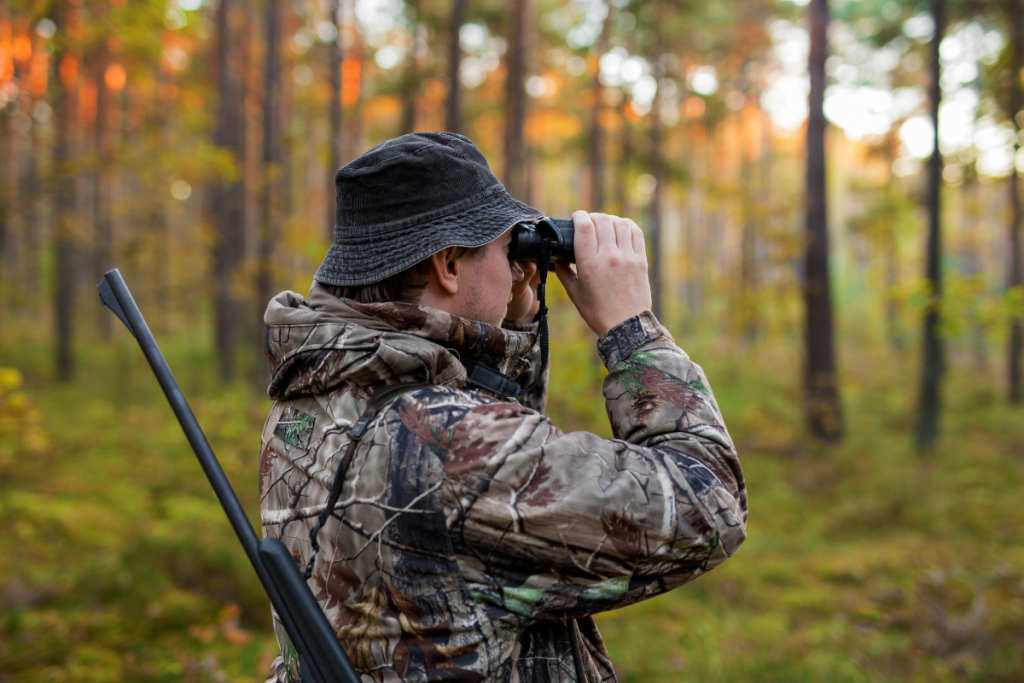 Hunter in camo jacket and bucket hat peers through binoculars in a autumn forest, rifle slung over his shoulder.
