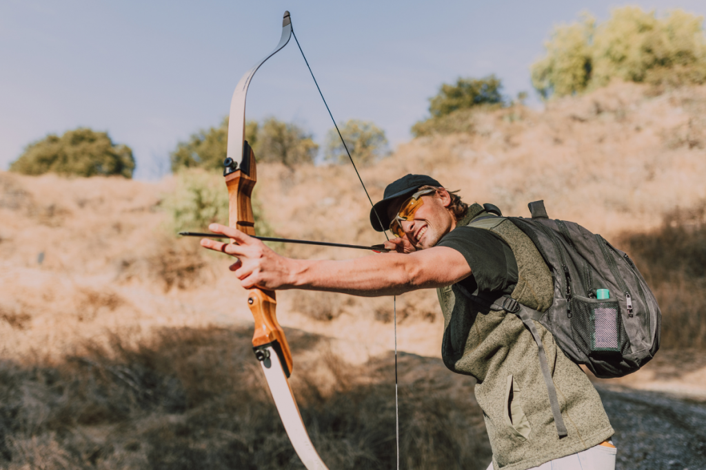 Archer outdoors drawing a wooden recurve bow with an arrow on a sunlit trail, wearing a cap and backpack.