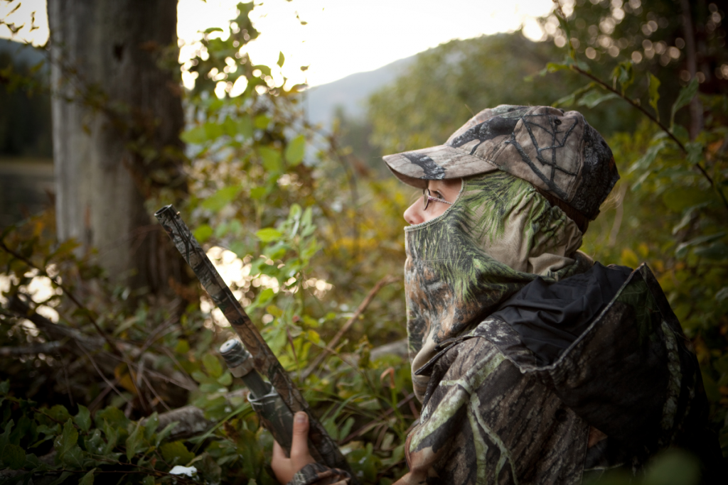 Person in camouflage clothing crouches among brush, holding a camouflaged shotgun, facing left in a forested area