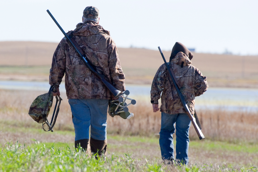 Two people in camo jackets walking away across a field, each carrying a shotgun and gear.