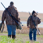 Two people in camo jackets walking away across a field, each carrying a shotgun and gear.