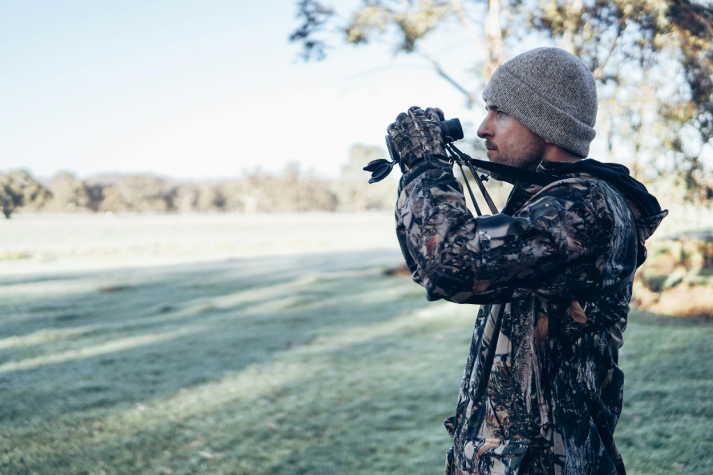 Man in camouflage jacket and beanie using binoculars in a grassy park or field, looking into the distance.