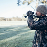 Man in camouflage jacket and beanie using binoculars in a grassy park or field, looking into the distance.